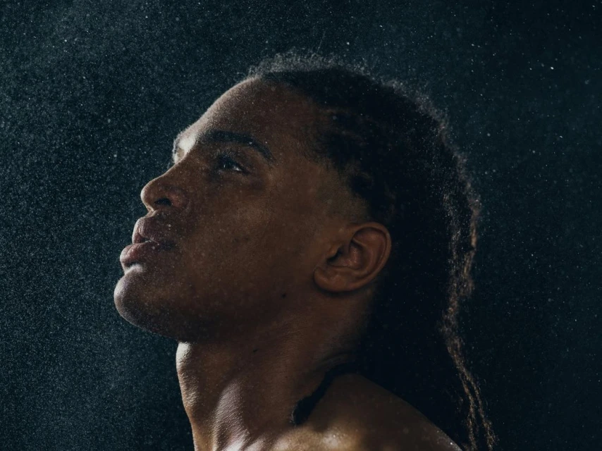 Profile shot of an adult male with dark skin and long dreadlocks, looking up while water droplets fall around him against a dark backdrop.