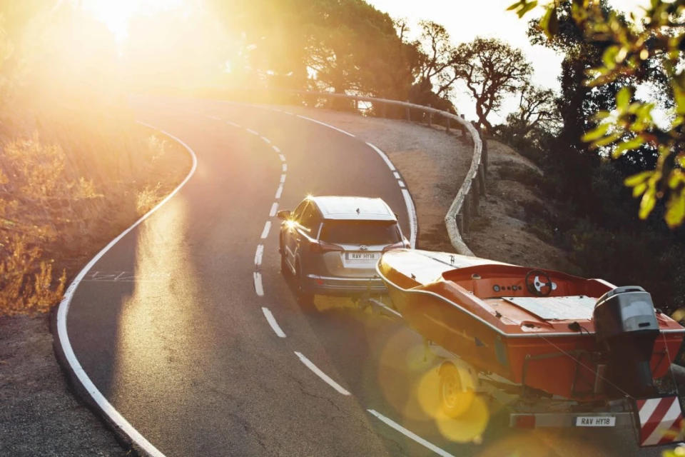 Car towing a boat on a winding road at sunset, surrounded by trees. The sunlight creates a warm, golden ambiance.