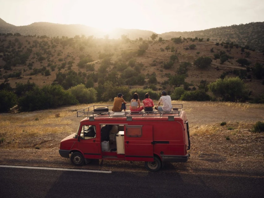Group of four people sits on the roof of a red van parked on an asphalt road against a backdrop of rolling hills and golden sunlight.