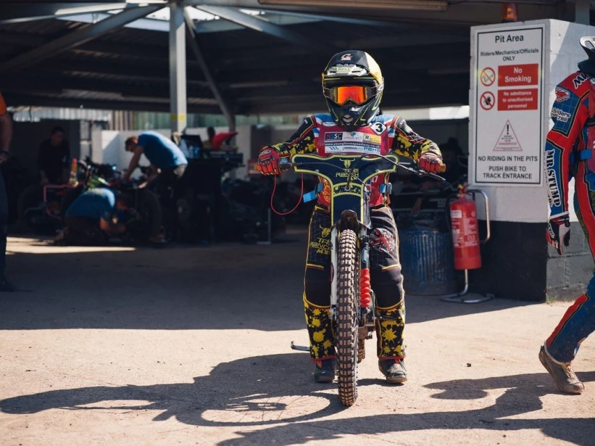 Motorcycle speedway racer in helmet and protective gear on bike at pit area, ready for race on sunny day, action sport competition.