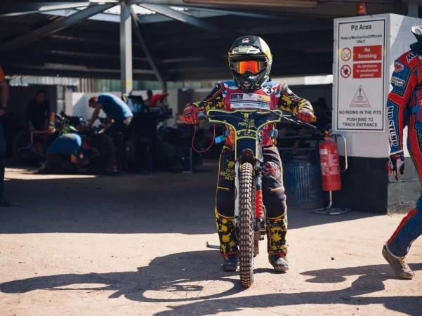 Motorcycle speedway racer in helmet and protective gear on bike at pit area, ready for race on sunny day, action sport competition.