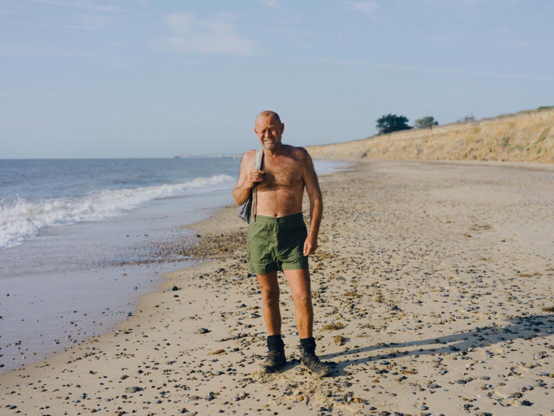 Elderly man wearing shorts standing on a sandy beach with ocean waves, carrying a bag over his shoulder, on a sunny day.