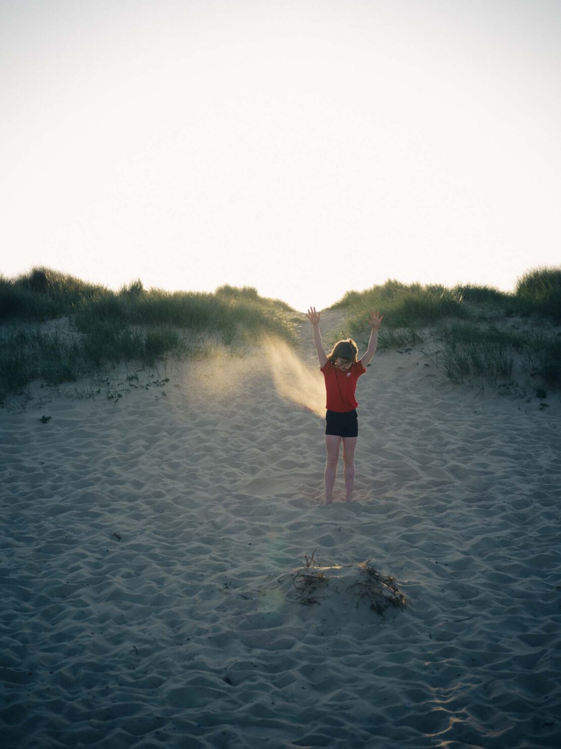 Child in red shirt stands on sand dune with arms raised at dusk, casting long shadows in a natural outdoor environment.