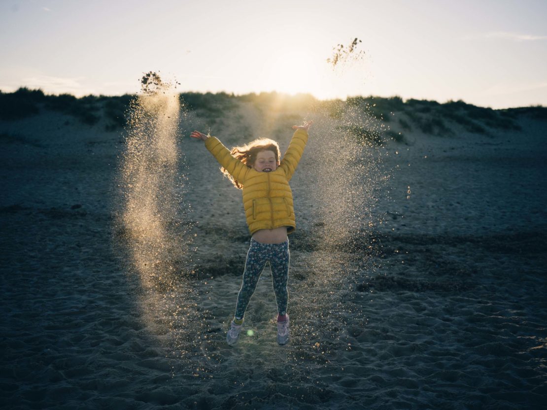 A happy young girl in a yellow puffer jacket jumps and throws sand on a beach at sunset with sand dunes in the background.