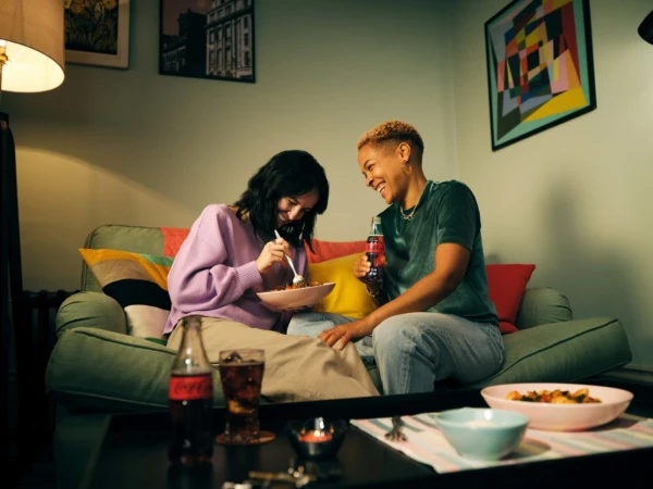 Two women enjoy a meal together on a green couch in a cozy living room, illuminated by lamp light, with drinks and artwork visible.