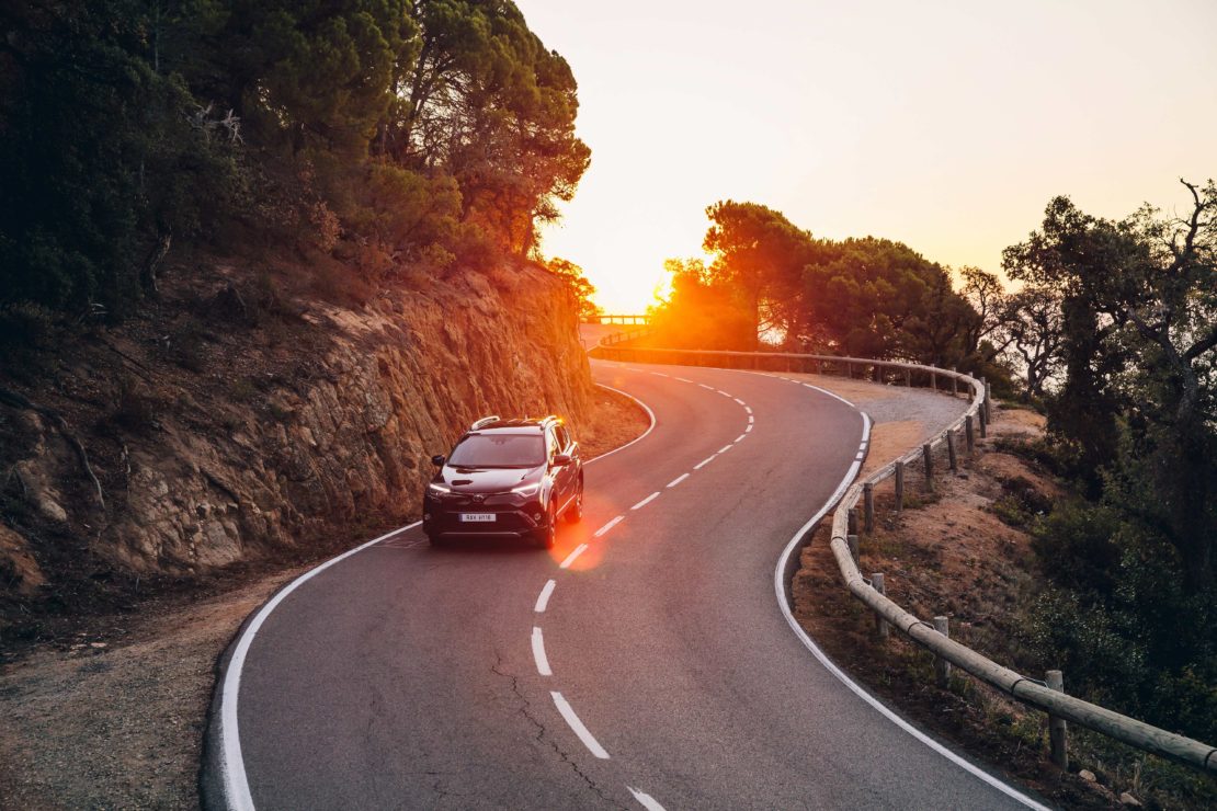 SUV driving on a winding mountain road at sunset. Vehicle navigates the curve with trees, rock formations, and a bright setting sun.
