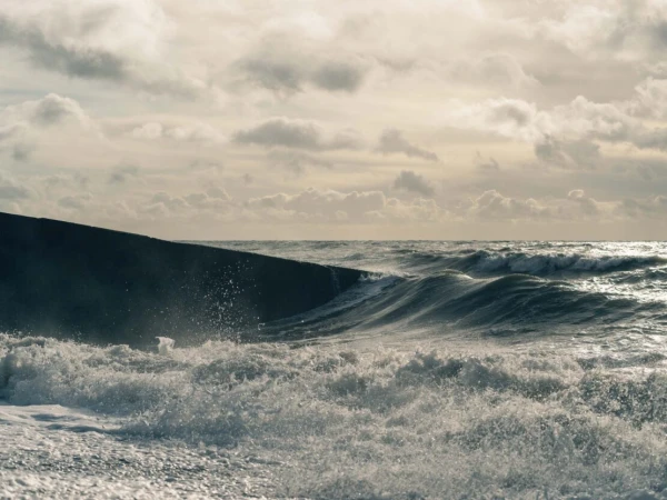 Choppy ocean waves crashing against a dark stone breakwater under a cloudy sky.