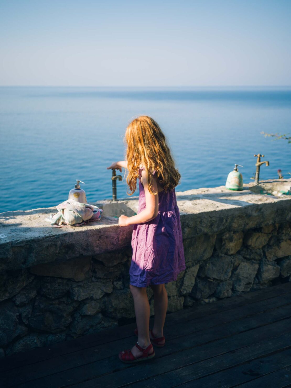 Child in a purple dress stands on a patio overlooking the ocean, turning on a water tap in an outdoor setting under a clear blue sky.
