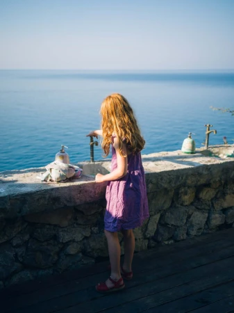 Child in a purple dress stands on a patio overlooking the ocean, turning on a water tap in an outdoor setting under a clear blue sky.