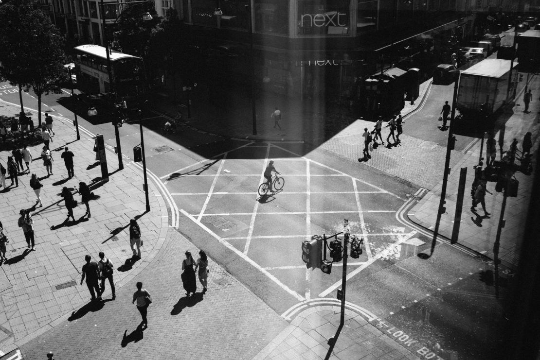 Black and white high-angle shot of a city intersection featuring pedestrians, a cyclist, traffic, and buildings, creating a dynamic urban scene.