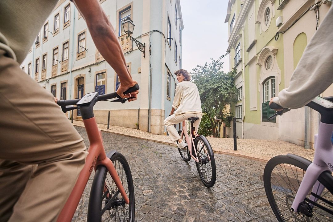 Three individuals ride modern electric bicycles on a cobblestone street lined with colorful buildings, creating a vibrant urban scene.