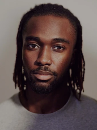 Close-up portrait of a Black man with dreadlocks and a nose stud wearing a grey t-shirt against a neutral background.