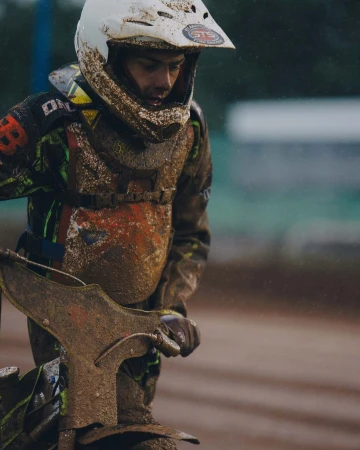 A mud-covered speedway racer stands with their motorcycle wearing full protective gear, including a white helmet and chest protector.