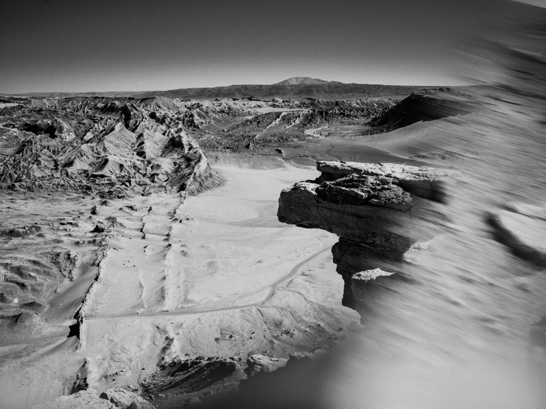 Monochrome landscape featuring a rocky desert terrain with cliffs and sand formations leading to distant mountains under a clear sky.
