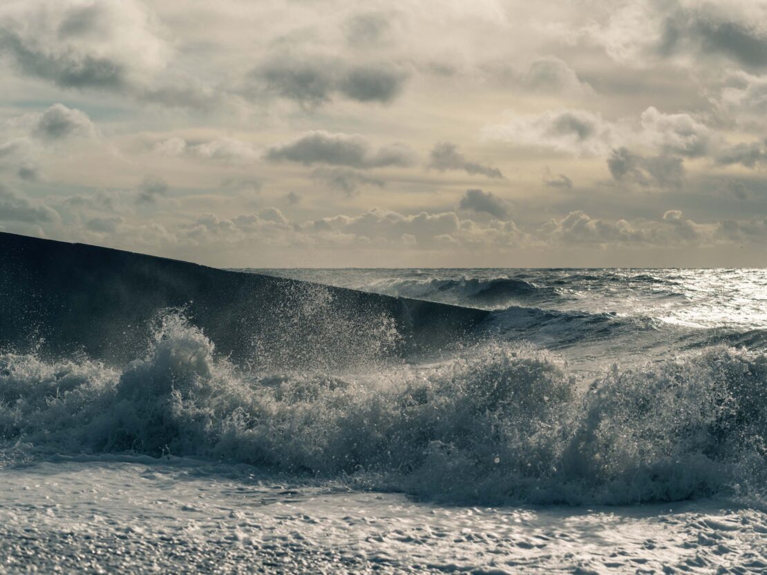 Waves crash against a sea wall under a cloudy sky, creating a dynamic coastal scene with textured water and diffused light.