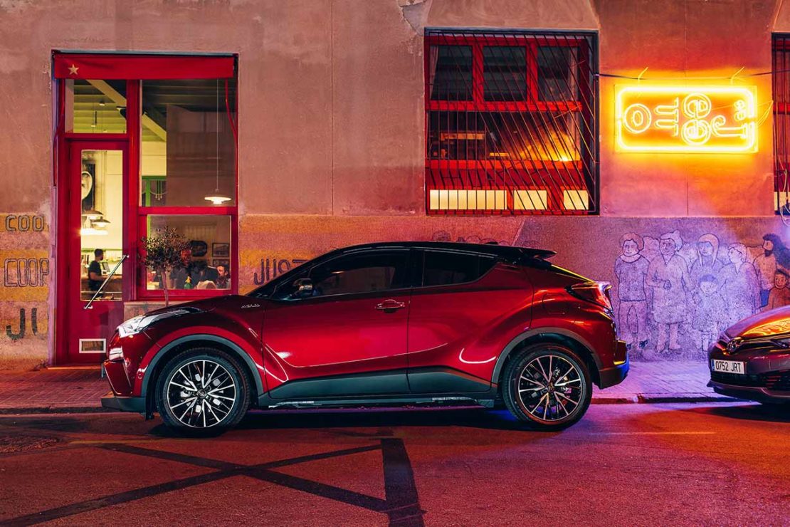 Maroon Toyota C-HR parked on a city street at night with a red neon sign glowing on the wall in the background next to a red doorway.
