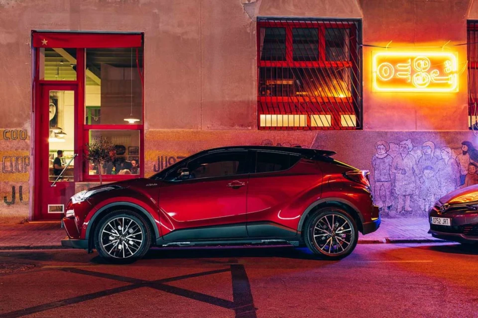 Maroon Toyota C-HR parked on a city street at night with a red neon sign glowing on the wall in the background next to a red doorway.