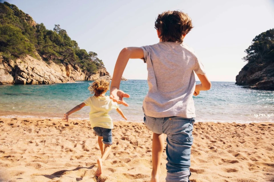 Two young boys run barefoot on a sandy beach towards the ocean on a sunny day. Trees and rock formations frame the tranquil cove.