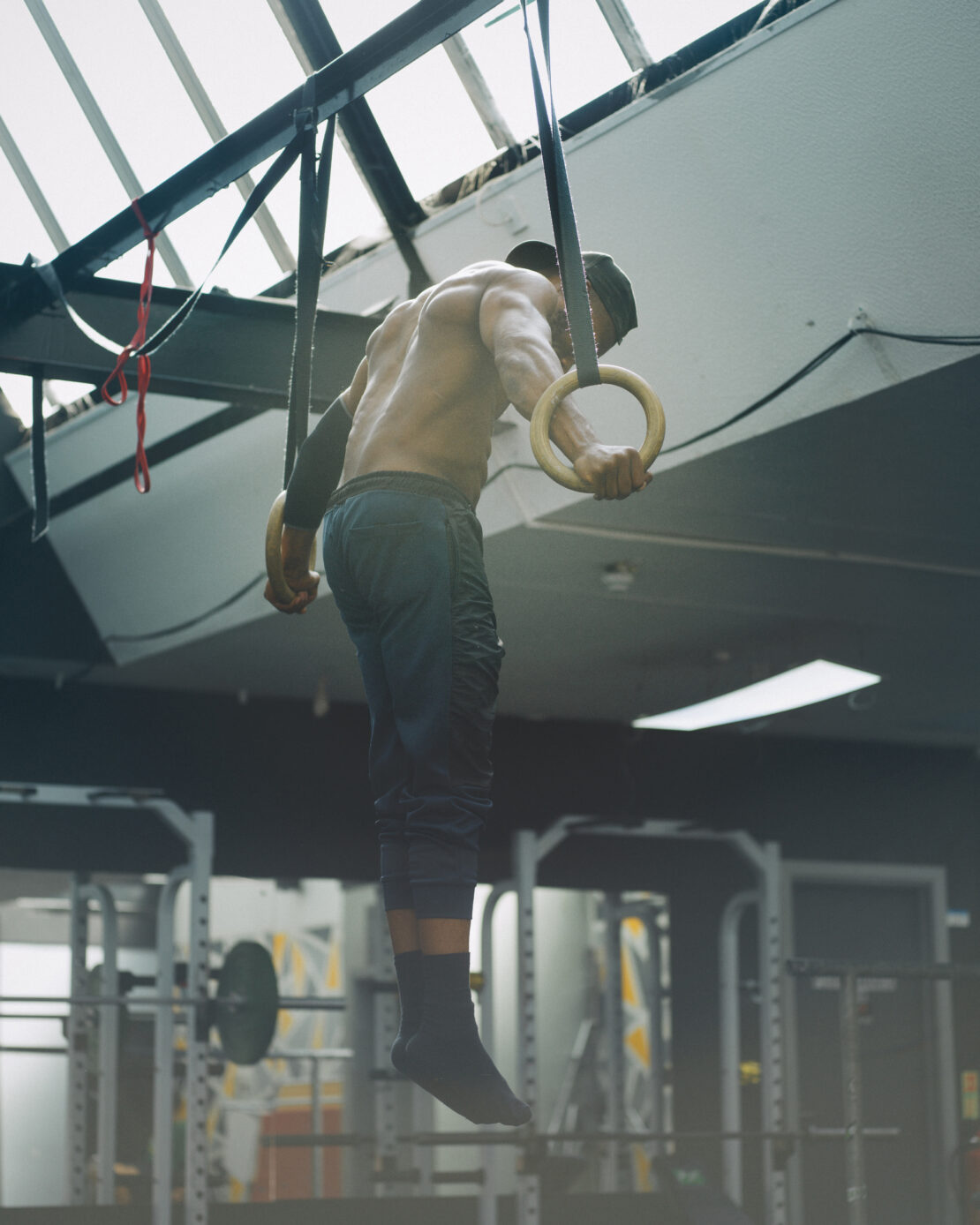 Athletic man performs gymnastic ring exercise in a gym. The man is shirtless with cap on, using wooden rings suspended from ceiling.
