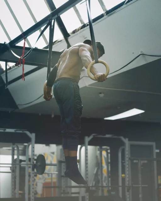 Athletic man performs gymnastic ring exercise in a gym. The man is shirtless with cap on, using wooden rings suspended from ceiling.