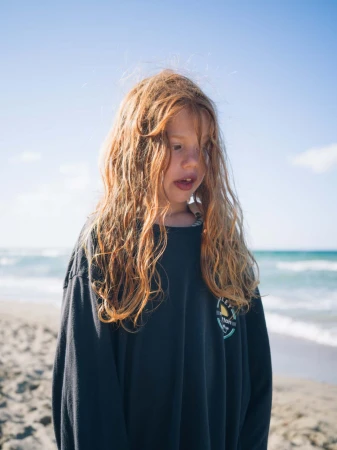 Young child with long curly red hair wearing a dark oversized long-sleeve shirt standing on a sandy beach near the ocean.