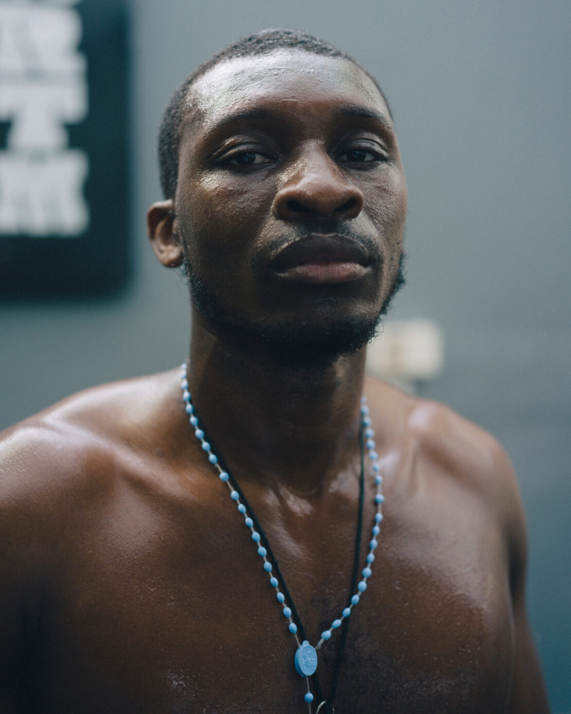 Close-up portrait of a muscular, sweaty, shirtless Black adult male looking intently at the camera with a blurred gray background.