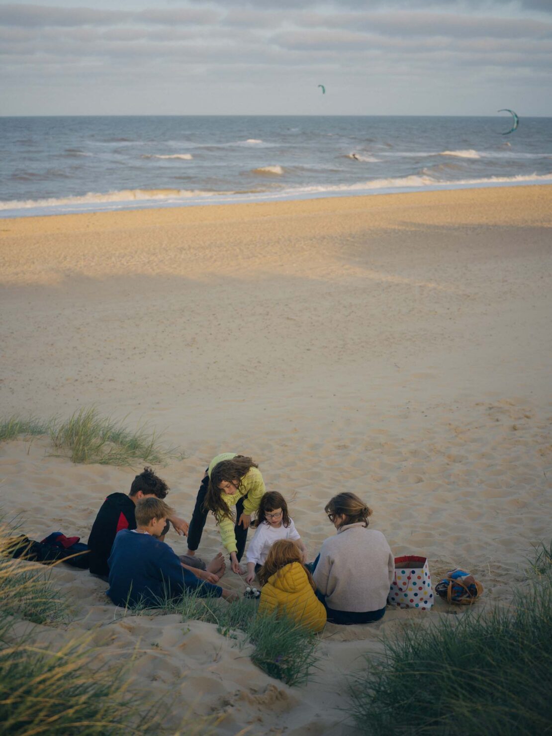 Group of people sitting on a sandy beach near the ocean with kitesurfers in the distance under a cloudy sky at dusk.
