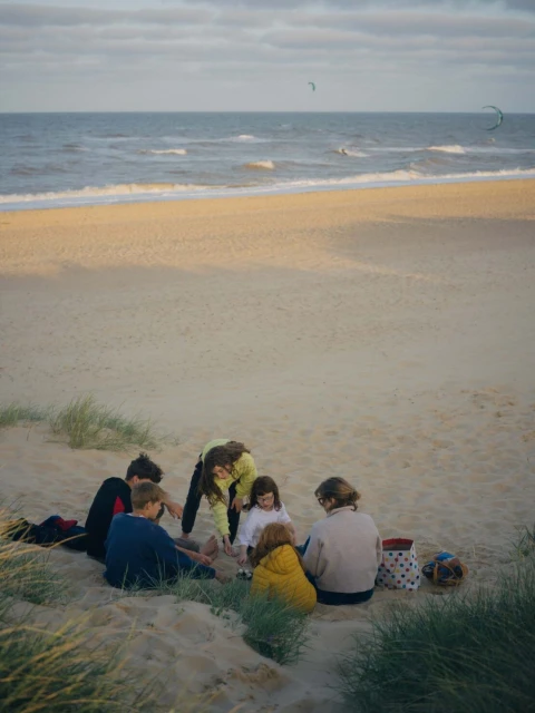 Group of people sitting on a sandy beach near the ocean with kitesurfers in the distance under a cloudy sky at dusk.