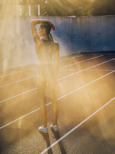 Athletic woman in sportswear stands on a running track with sunlight, wearing a sports bra, shorts, and sneakers, in an outdoor setting.