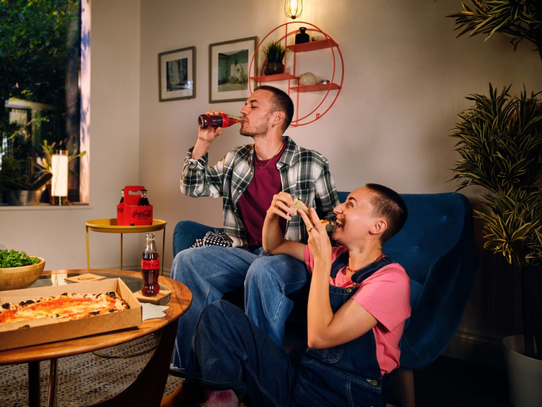 Couple enjoying Coca-Cola and pizza in a cozy living room. Two people are relaxing on a sofa, with snacks and drinks on a nearby table.