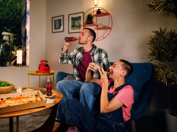 Couple enjoying Coca-Cola and pizza in a cozy living room. Two people are relaxing on a sofa, with snacks and drinks on a nearby table.