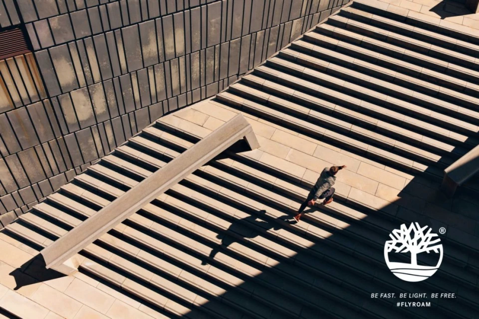 High-angle view of a person walking up concrete stairs next to a concrete bench; long shadows and bright sun with Timberland logo.