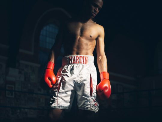 An athletic African-American male boxer wearing red boxing gloves and white boxing shorts stands in a boxing ring in a dimly lit environment.