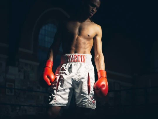 An athletic African-American male boxer wearing red boxing gloves and white boxing shorts stands in a boxing ring in a dimly lit environment.