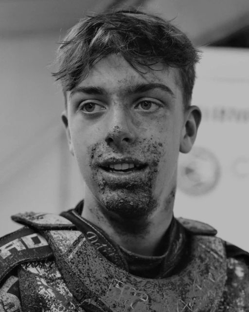 Black and white portrait of a young man with mud on his face and sporting protective gear, looking off to the side.