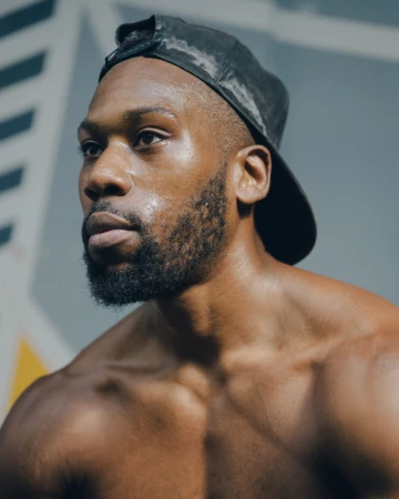 Close-up portrait of a sweaty, muscular, shirtless Black male athlete with a beard wearing a black baseball cap, looking off-camera.