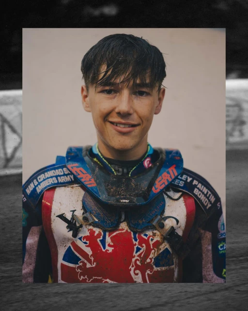 Close-up portrait of a smiling young adult male athlete wearing a protective racing suit with logo details, presented as a photograph.