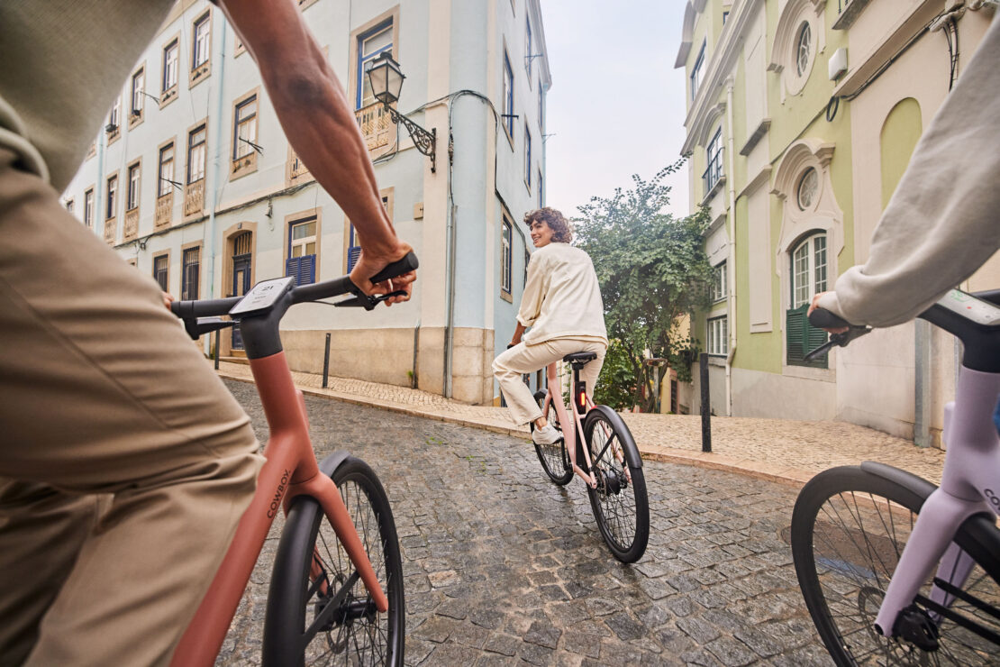 Three people riding electric bikes down a cobblestone street lined with European-style buildings on an overcast day in an urban setting.