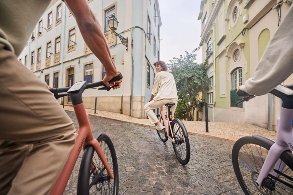 Three people riding electric bikes down a cobblestone street lined with European-style buildings on an overcast day in an urban setting.