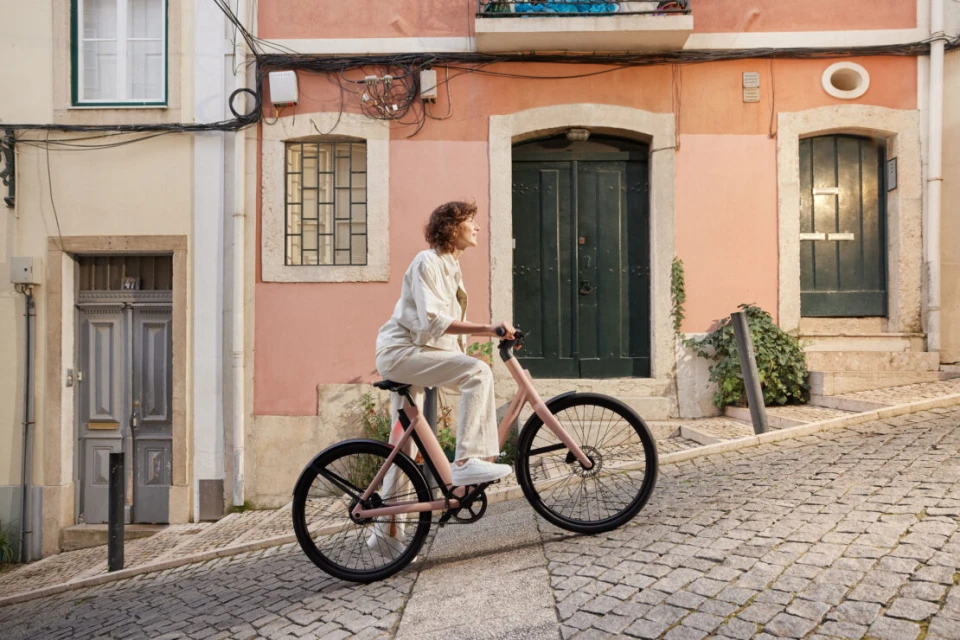 Woman in white outfit riding a pink bicycle on a cobblestone street with colorful buildings in the background on a sunny day.