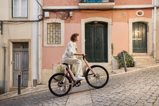 Woman in white outfit riding a pink bicycle on a cobblestone street with colorful buildings in the background on a sunny day.