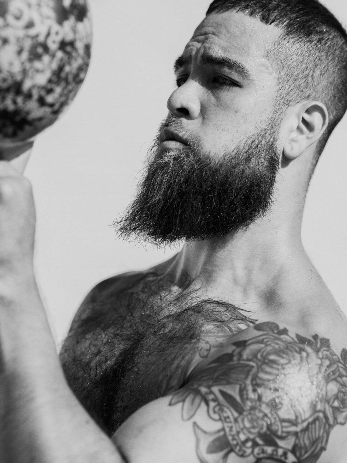 Monochrome portrait of an adult male with a beard, shoulder tattoo and a ball. Head and shoulders, neutral expression, white background.