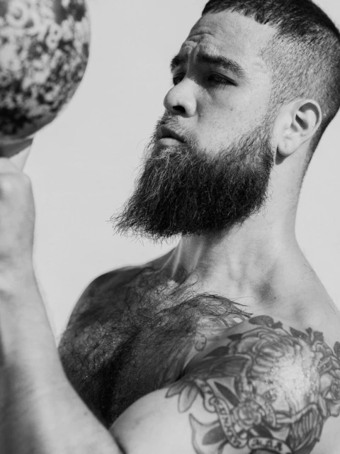 Monochrome portrait of an adult male with a beard, shoulder tattoo and a ball. Head and shoulders, neutral expression, white background.