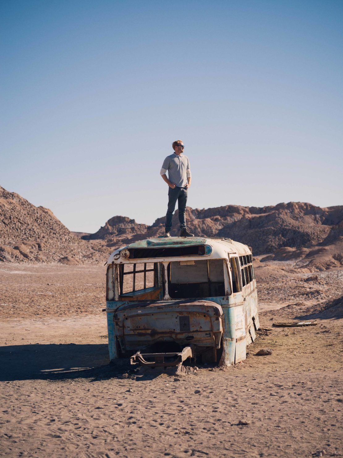 Adult male stands on rusted bus in desert landscape under clear sky; remote travel adventure or post-apocalyptic abandoned vehicle concept.