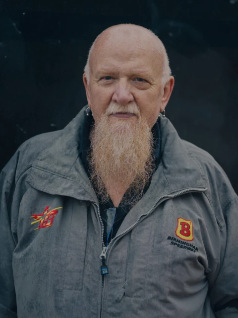 Portrait of an older man with a long beard, wearing a racing jacket, posing against a dark background in a medium shot.