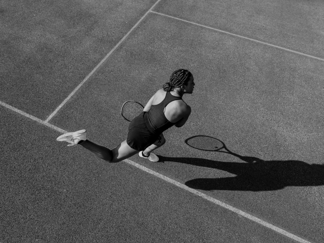 Black and white high-angle shot of an adult woman playing tennis on a court, wearing a tank top, skirt, and athletic shoes.