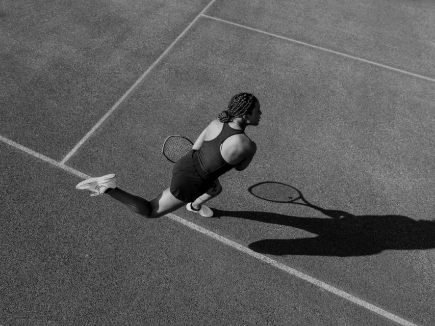 Black and white high-angle shot of an adult woman playing tennis on a court, wearing a tank top, skirt, and athletic shoes.