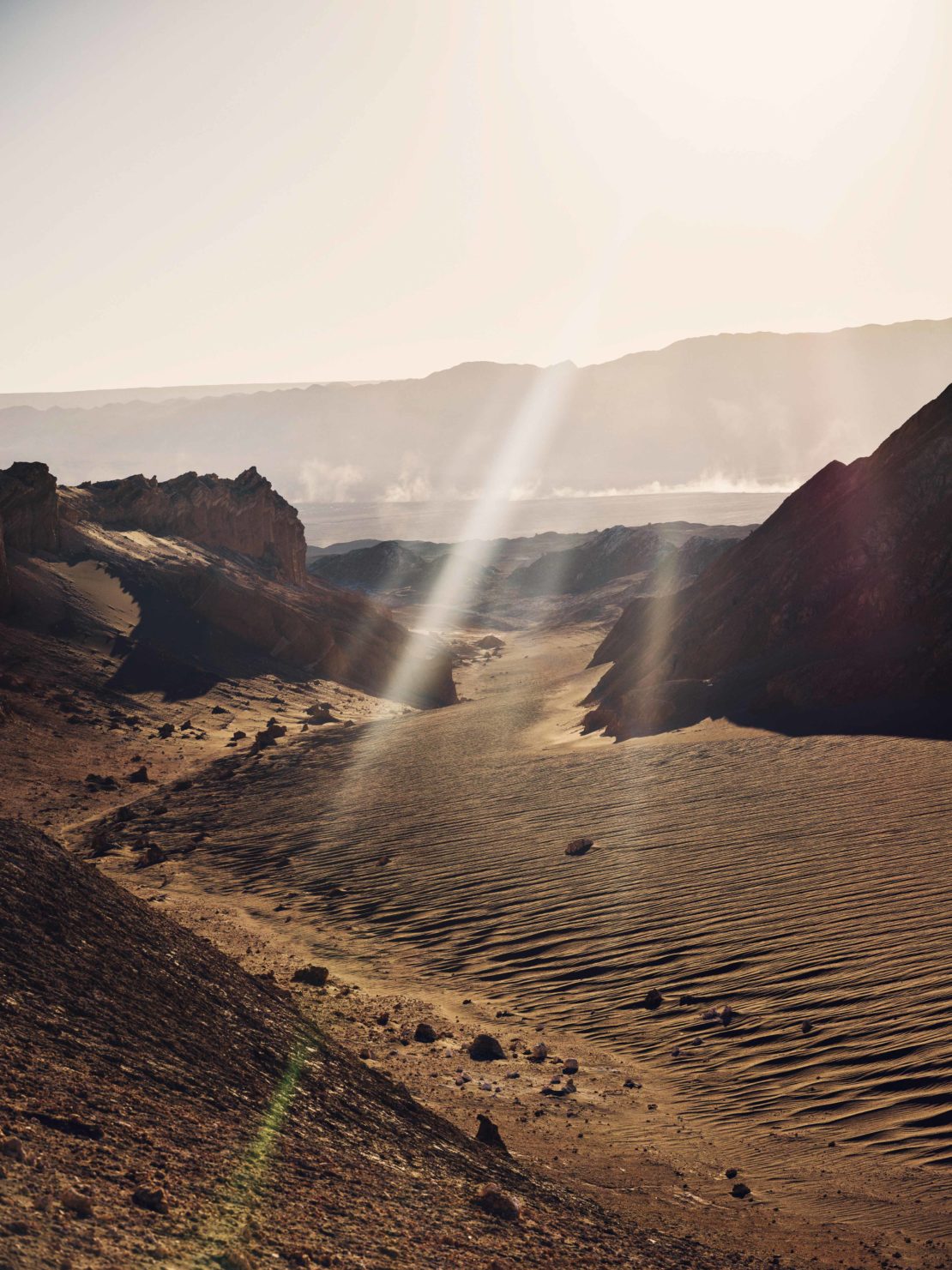 Desert landscape view with bright sunlight creating lens flares over rocky terrain, distant hills and clear sky.