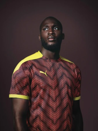 Black man wearing a patterned red and black jersey with yellow shoulder panels against a dark background