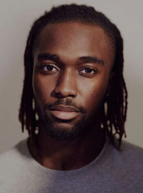 Portrait of a young Black man with dreadlocks, wearing a gray t-shirt, against a light neutral background, focusing on his facial features.